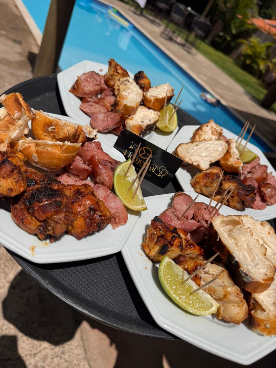 Pratos com carnes grelhadas, linguiça, pão e limão servidos à beira da piscina